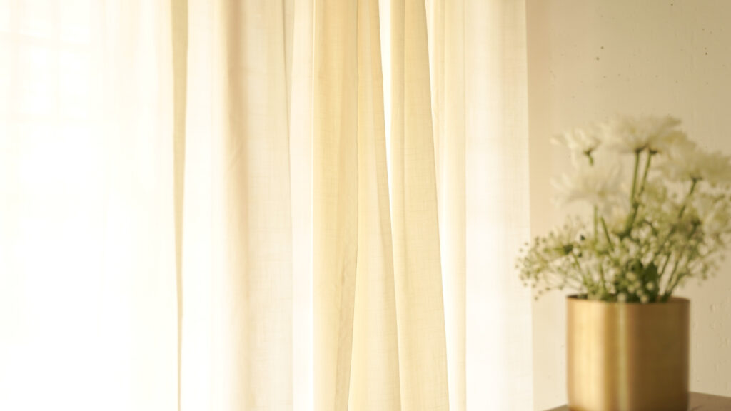 Sheer ivory cotton curtains letting in soft daylight beside a brass vase of white flowers